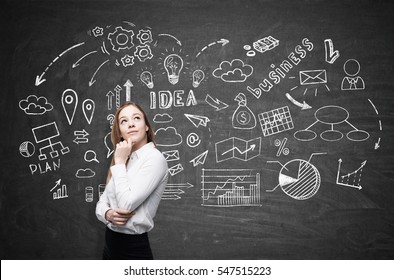 Portrait Of A Pondering Blond Girl Standing Near A Blackboard With A Business Idea Sketch Depicted On It. Concept Of Young Entrepreneurship.