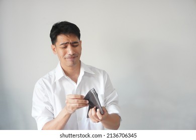 Portrait of poor Asian guy looks at his empty wallet, unhappy, worried, lose money, funny facial expressions. Concept of employee, businessman, office worker, bankruptcy, and unemployment in crisis