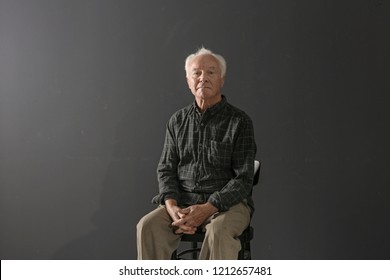 Portrait Of Poor Elderly Man Sitting On Chair Against Dark Background