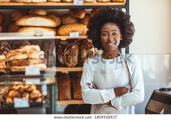 Portrait of positive African American young woman\
working in own Bakery shop, looking at camera with toothy smile.\
Pretty baker smiling at camera. Small Bakery shop owner standing in\
front of store