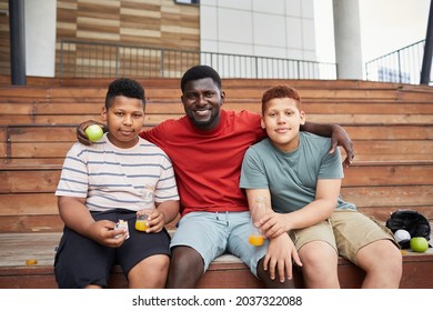 Portrait Of Positive African-American Father Sitting On Wooden Bleacher Bench And Embracing Sons At Sports Match