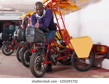 Portrait of positive African-American man bikecab driver standing near rickshaw cycle