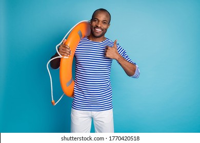 Portrait of positive afro american guy hold circle lifeguard buoy approve life sand safety quality show thumb up sign wear sailor striped shirt white shorts isolated blue color background