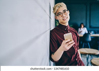 Portrait Of Positive Asian Millennial Male Blogger In Optical Spectacles For Provide Eyes Protection Holding Modern Cellular Phone In Hand And Smiling At Camera Standing Indoors.Hipster Guy With Phone