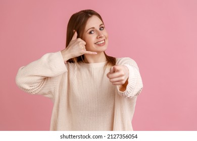 Portrait Of Positive Attractive Blond Woman Showing Telephone Hand Gesture Near Head, Smiling And Pointing To Camera, Wearing White Sweater. Indoor Studio Shot Isolated On Pink Background.