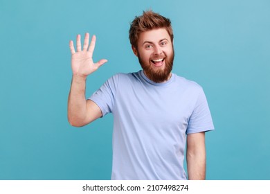 Portrait Of Positive Bearded Man Greeting You Rising Hand And Waving, Saying Hi, Glad To See You, Looking At Camera With Toothy Smile. Indoor Studio Shot Isolated On Blue Background.