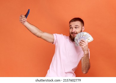 Portrait of positive bearded man making selfie photo with bunch of dollars on smartphone, bragging with wealth, wearing pink T-shirt. Indoor studio shot isolated on orange background. - Shutterstock ID 2157602779