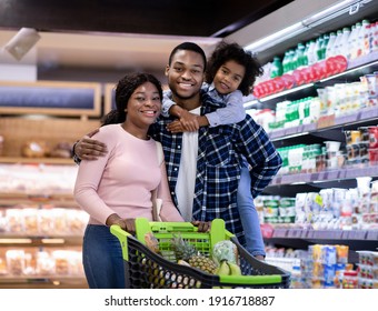Portrait of positive black family with shopping cart smiling and looking at camera at modern supermarket. Young parents and their cute little daughter buying groceries at huge mall