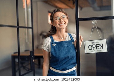 Portrait Of Positive Business Woman Standing At Cafeteria Door Entrance. Cheerful Young Waitress In Blue Apron Near Glass Door With Open Signboard And Looking At Camera. Excited Small Business Owner.