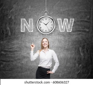 Portrait of a positive businesswoman standing near a blackboard with a clock and a word now on it. Concept of time to act.