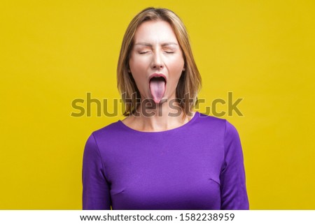 Portrait of positive carefree woman with fair hair in purple dress standing with closed eyes and demonstrating tongue, naughty disobedient expression. indoor studio shot isolated on yellow background