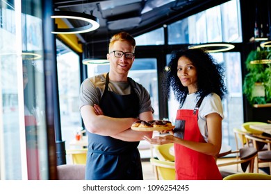 Portrait of positive caucasian barista with crossed hands teamworking with cheerful african american female confectioner which holding plate with tasty delicious pastries standing in common bakery