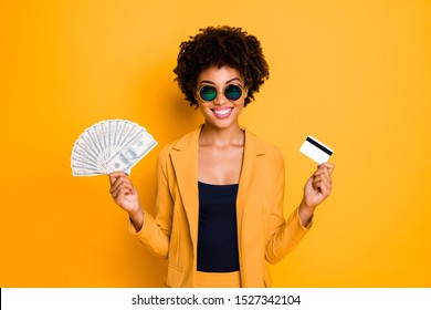 Portrait of positive cheerful brown curly hair afro american girl hold money fan debit card got cashback profit want buy pay on sales discounts wear style jacket suit isolated yellow color background