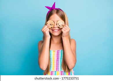 Portrait of positive cheerful child girl hold chocolate cookies close cover eyes face enjoy snack wear shine trendy stylish skirt isolated over blue color background