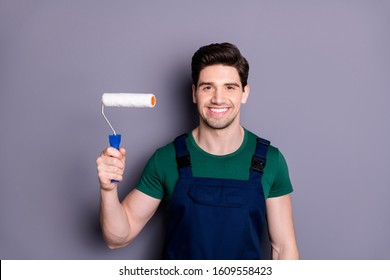 Portrait Of Positive Cheerful House Painter Man Hold White Roller Ready To Change His House Wear Green T-shirt Blue Uniform Isolated Over Grey Color Background