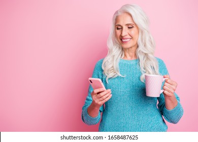 Portrait of positive cheerful modern old woman granny use smartphone follow social media post comment hold hot latte beverage mug wear blue jumper isolated over pink color background