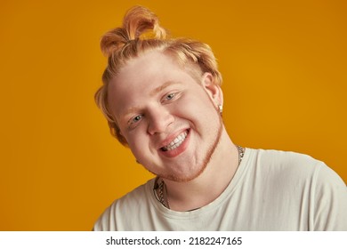 Portrait Of Positive Chubby Red-haired Guy With A High Ponytail Dressed In A White T-shirt Joyfully Smiling. Studio Shot On A Yellow Background. Modern Young People. Emotions.