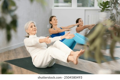 Portrait of positive concentrated elderly woman doing Havasana pose to strengthen abs and core muscles during group yoga session - Powered by Shutterstock