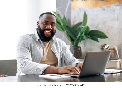 Portrait Of A Positive Confident Successful African American Young Man, Sales Manager, Ceo Or Freelancer, Sits At A Work Desk With Laptop In Modern Office, Looking At The Camera, Smiles Friendly