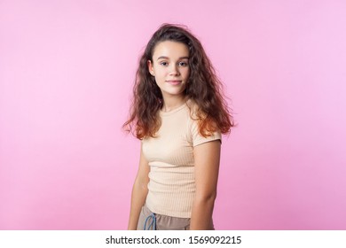 Portrait of positive confident teen girl with long curly brunette hair wearing casual style beige clothes looking at camera with happy calm expression. indoor studio shot isolated on pink background