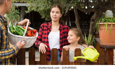 Portrait Of Positive Couple Of Gardeners With Daughter Standing In Garden Near Wooden Fence With Gathered Vegetables Talking About Rich Harvest