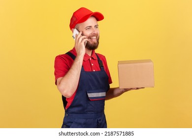 Portrait Of Positive Courier Woman Standing Holding Cardboard Box, Parcel For Client And Talking Via Cell Phone, Wearing Overalls And Red Cap. Indoor Studio Shot Isolated On Yellow Background.