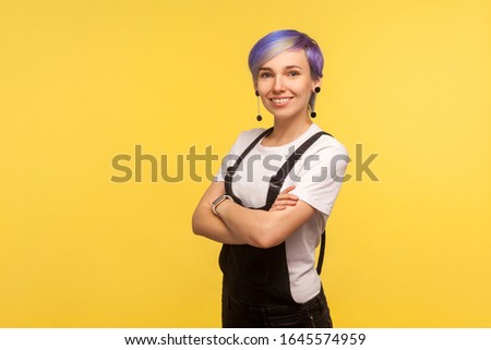 Portrait of positive cute hipster woman with violet dyed short hair in denim overalls smiling at camera and crossing hands, stylish fashionable appearance. isolated on yellow background, studio shot