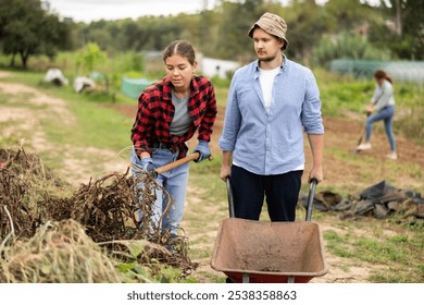 Portrait of positive employees cleaning backyard. Woman and man takes out bunch of branches, puts garbage in wheelbarrow, and cleans up next to cottage and ranch. - Powered by Shutterstock