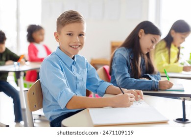 Portrait of positive European schoolboy sitting at his desk in classroom during lesson in secondary school and smiling at camera - Powered by Shutterstock