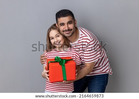 Portrait of positive family with red present box in hands, father and daughter giving gift for mother's birthday, expressing happiness. Indoor studio shot isolated on gray background.