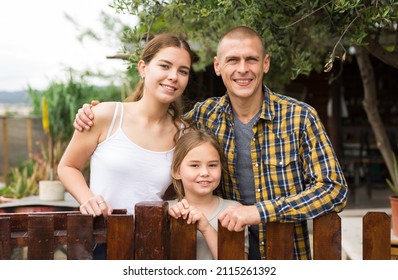 Portrait Of Positive Family Of Three Posing Near Wooden Fence In Backyard Garden, Family Gardening Concept