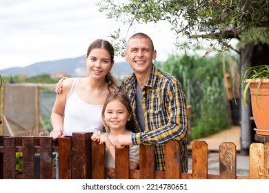 Portrait Of Positive Family Of Three Posing Near Wooden Fence In Backyard Garden, Family Gardening Concept