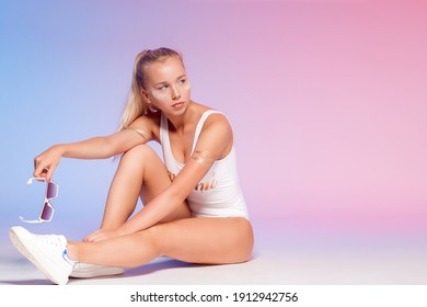 Portrait Of Positive Fashion Young Girl In A White Swimsuit And Stylish Sunglasses In Studio At Pastel Colors Background. Model Looking At Right.