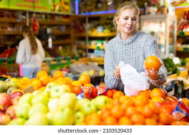 Portrait Of Positive Female Shopper Making Purchases In Supermarket, Looking For Fresh Sweet Oranges ..