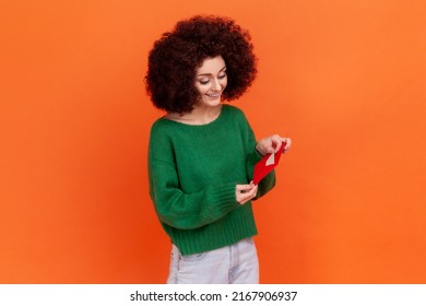 Portrait Of Positive Female Woman With Afro Hairstyle Wearing Green Casual Style Sweater Standing Holding Red Envelope, Reading Romantic Letter. Indoor Studio Shot Isolated On Orange Background.