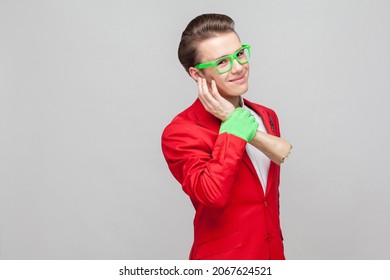 Portrait Of Positive Funny Gentleman With Eyeglasses In Red Tuxedo And Green Gloves Holding Zombie Hand, Stroking His Cheek Or Pretending To Make Phone Call. Studio Shot Isolated On Gray Background