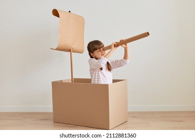 Portrait of positive funny little girl sitting in cardboard box with paper flag, playing ship captain, pretending she is in the sea, holding big spotting scope, looking far.