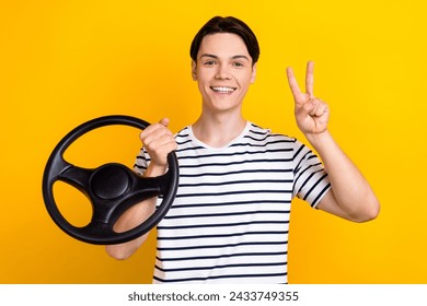 Portrait of positive guy with brunet hair wear stylish t-shirt hold steering wheel showing v-sign isolated on yellow color background