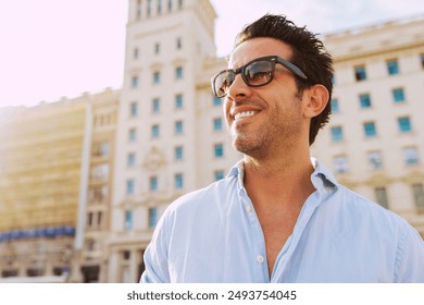 Portrait of a positive handsome businessman wears shirt and sunglasses, broadly smiles looking away, standing in front of a building at city during sunset. - Powered by Shutterstock