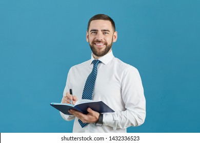 Portrait Positive Handsome Young Business Assistant With Beard Standing Against Blue Background And Making Notes In Diary