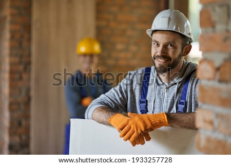 Portrait of positive, handsome young male builder in hard hat smiling at camera, holding drywall while working at construction site