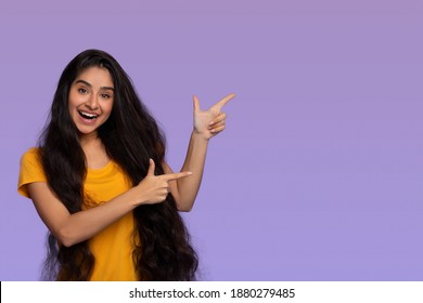 Portrait Of Positive Happy Indian Lady Pointing Two Fingers Aside At Free Copy Space Advertising Your Text, Standing Isolated Over Pastel Purple Studio Background. Emotional Woman With Long Dark Hair