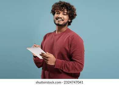 Portrait of positive Indian man with curly hair holding notebook and pen, writing looking at camera, isolated on blue background. Student studying, exam preparation, successful education - Shutterstock ID 2324747041