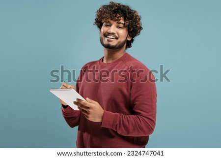 Portrait of positive Indian man with curly hair holding notebook and pen, writing looking at camera, isolated on blue background. Student studying, exam preparation, successful education