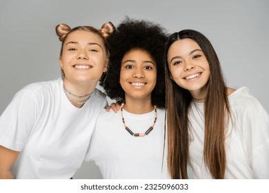 Portrait of positive interracial girlfriends in white t-shirts hugging african american friend and looking at camera together isolated on grey, teenage friends having fun together - Shutterstock ID 2325096033