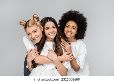 Portrait of positive and interracial teenage girls in white t-shirts hugging brunette girlfriend and looking at camera isolated on grey, adolescence models and generation z concept - Powered by Shutterstock