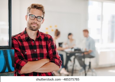 Portrait of a positive looking young business professional standing with his arms crossed with coworkers talking in the background. - Powered by Shutterstock