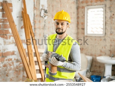 Portrait of positive man builder posing with handheld demolition hammer at indoor construction site