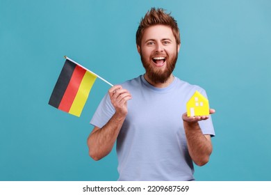 Portrait Of Positive Optimistic Handsome Bearded Man Holding Toy Paper House And Flag Of Germany In Hand, Moving Into Another Country. Indoor Studio Shot Isolated On Blue Background.