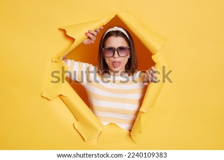 Portrait of positive optimistic woman wearing striped T-shirt, hair band and sunglasses, breaking through paper hole in yellow wall, looking at camera, showing tongue out.